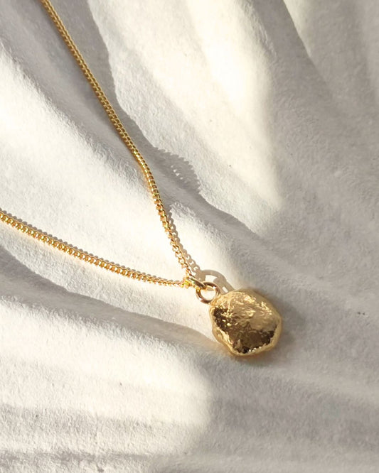 Molten gold pebble pendant on delicate chain lying in sunshine on white ceramic shell dish