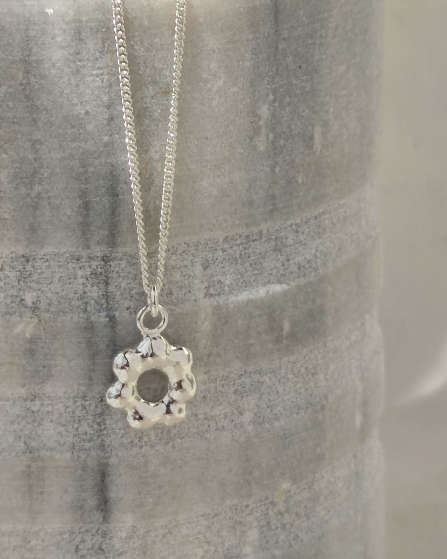 Small flower-shaped molten silver pendant hanging from fine sterling silver chain in front of grey stone vessel