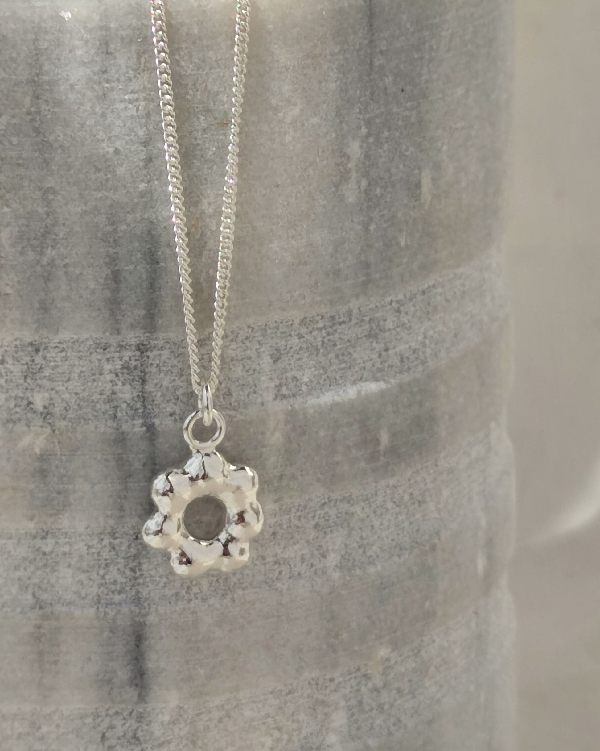 Small flower-shaped molten silver pendant hanging from fine sterling silver chain in front of grey stone vessel