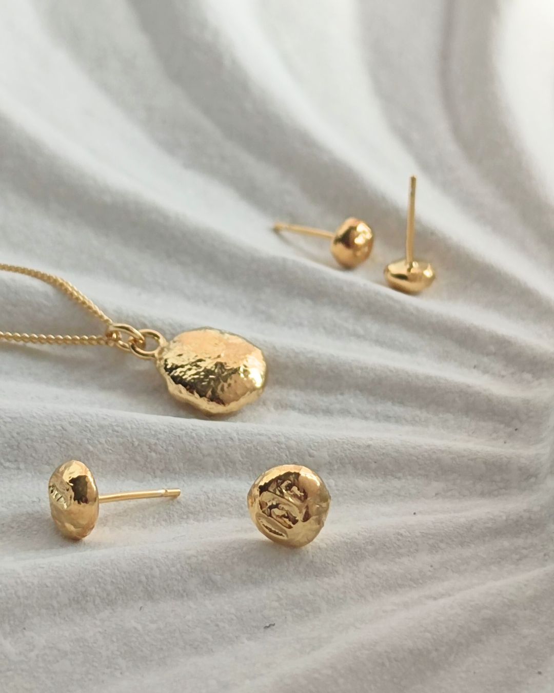 Handmade molten gold pendant and stud earrings lying on textured white ceramic shell dish