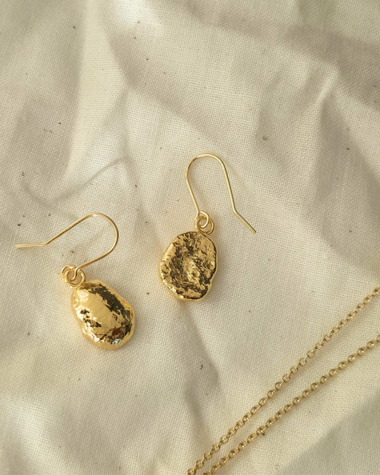 Gold oval shaped earrings with a textured surface lying on a beige fabric background