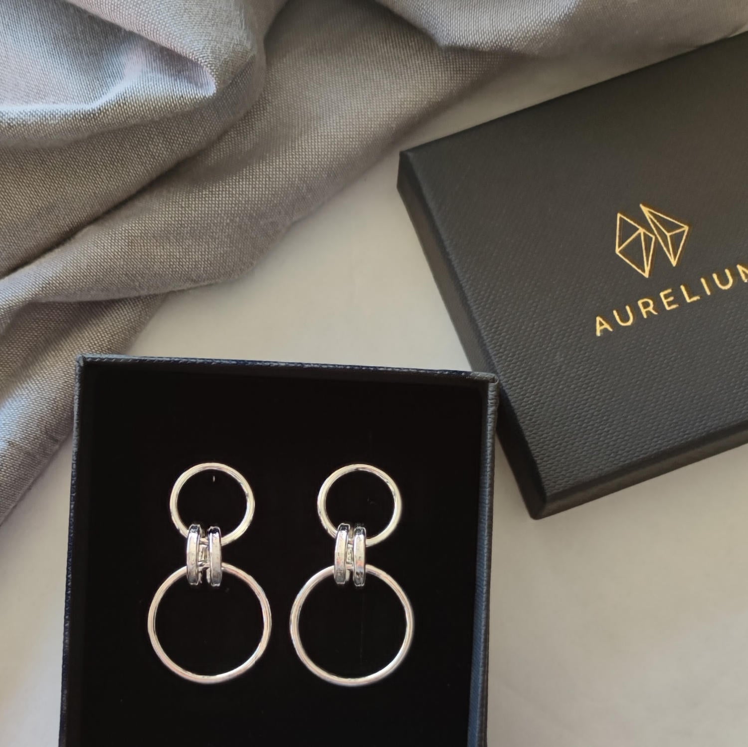 Handmade silver drop earrings created from small and large loops and presented in open black gift box beside lid printed with gold logo and "Aurelium" in gold lettering