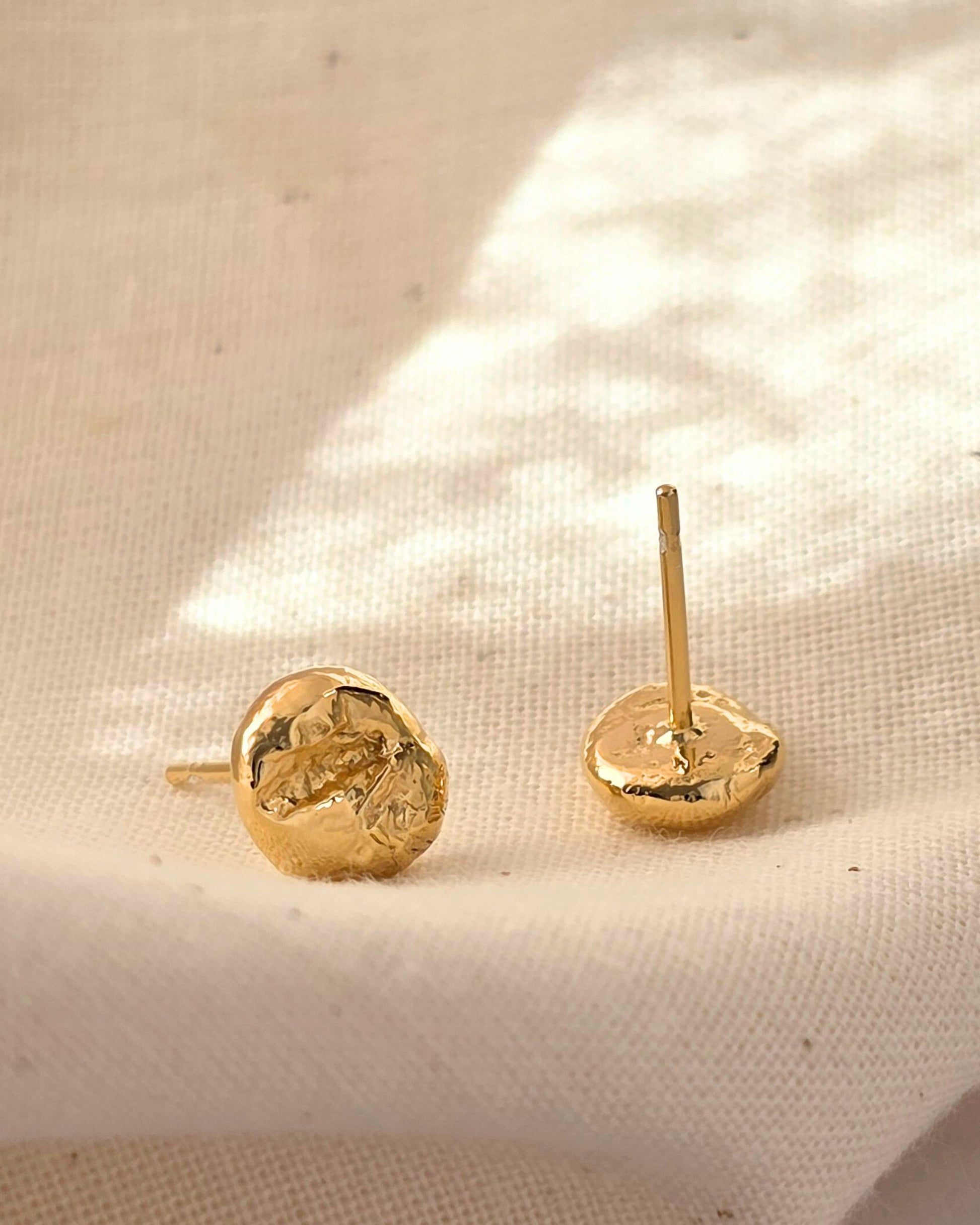 Small nugget-shaped gold stud earrings with a rippled texture lying in the sunshine on beige fabric