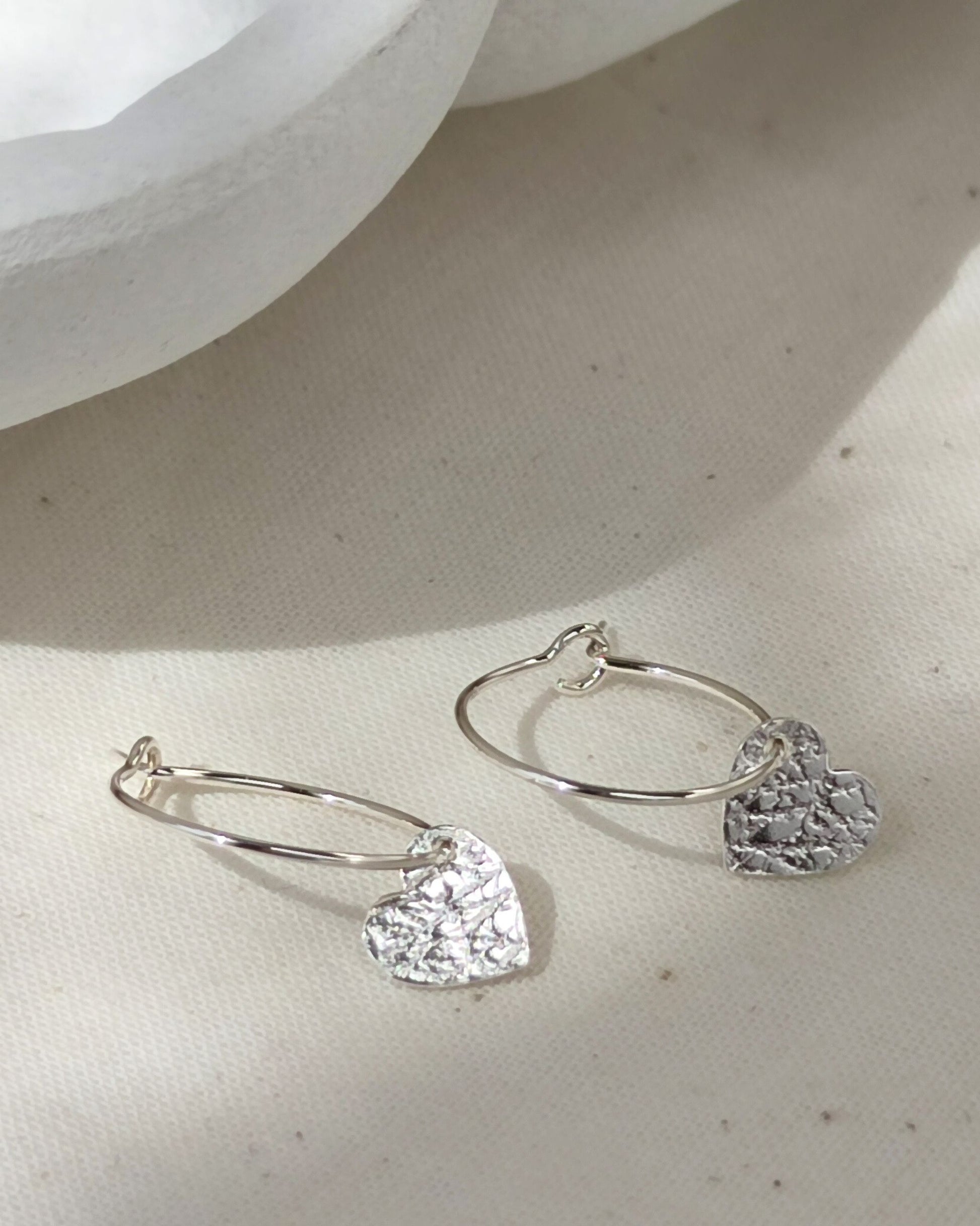 Small sterling silver hearts with delicate texture on silver hoops lying on cream fabric with white ceramic dish in background