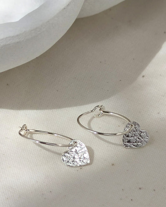 Small sterling silver hearts with delicate texture on silver hoops lying on cream fabric with white ceramic dish in background