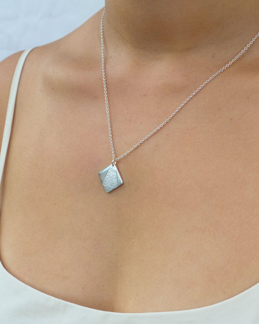 Person wearing small silver square pendant with partially textured surface in triangle shape hanging from fine silver chain