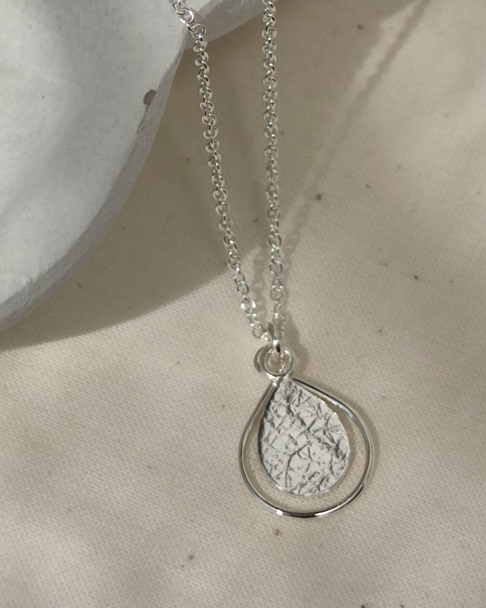 Sterling silver pendant consisting of textured teardrop framed within thin wire teardrop and hanging on fine sterling silver cable chain, lying on beige fabric