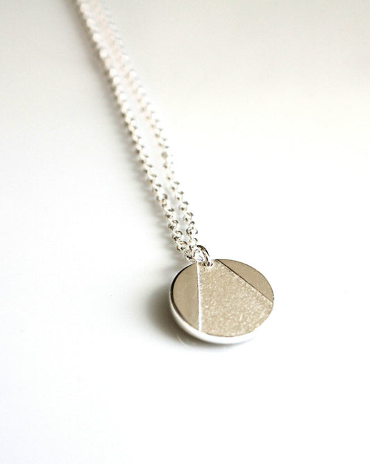 Circle-shaped silver pendant with partially textured triangle design hanging from thin silver chain on white background