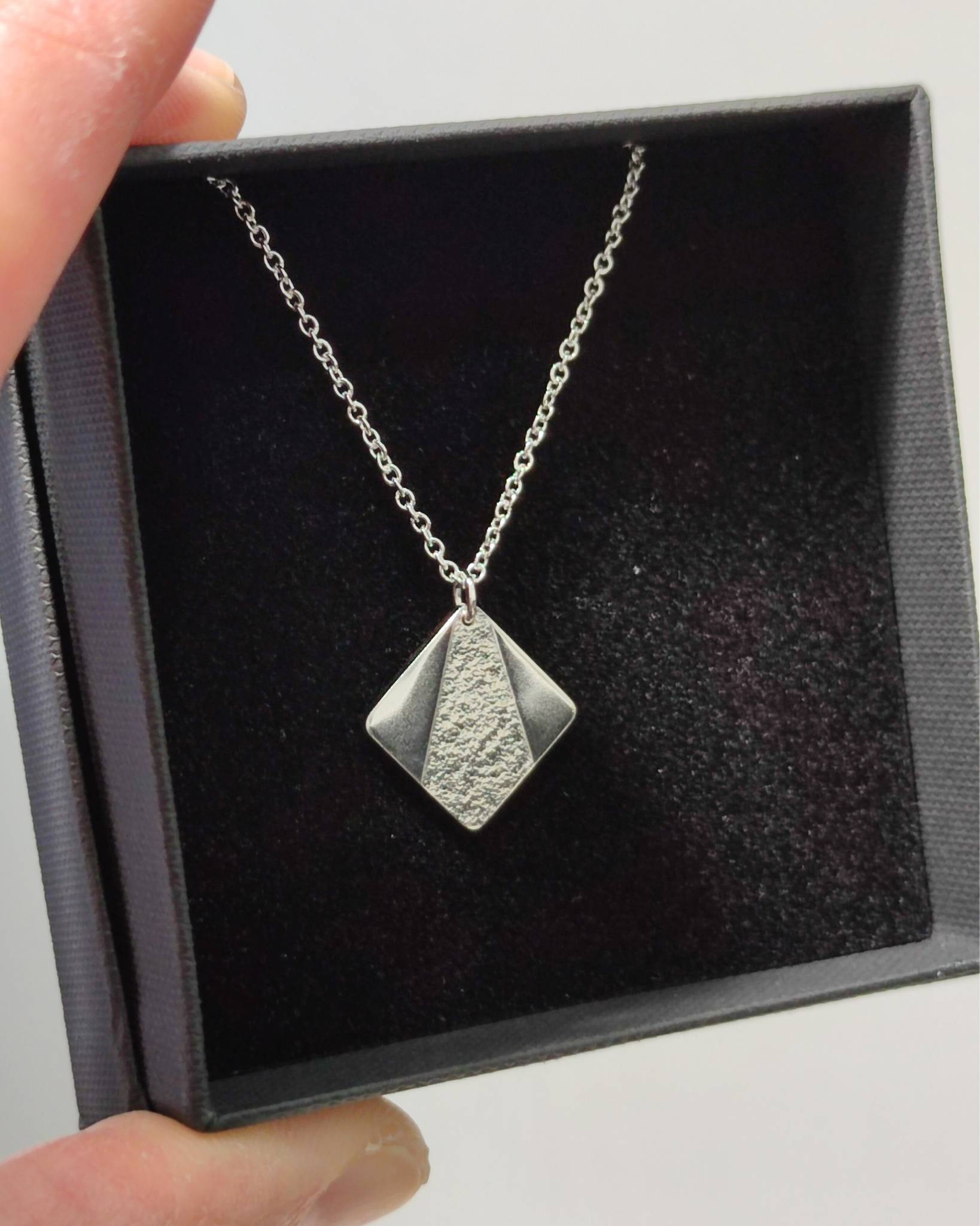 Hand holding a black jewellery gift box containing a small silver square pendant with partially textured triangle pattern