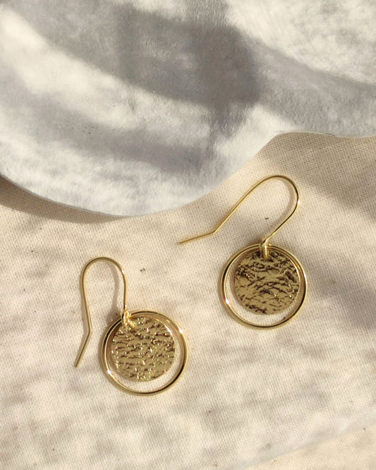 Handmade gold textured circle earrings framed with thin gold loops lying in sunshine beside white ceramic dish