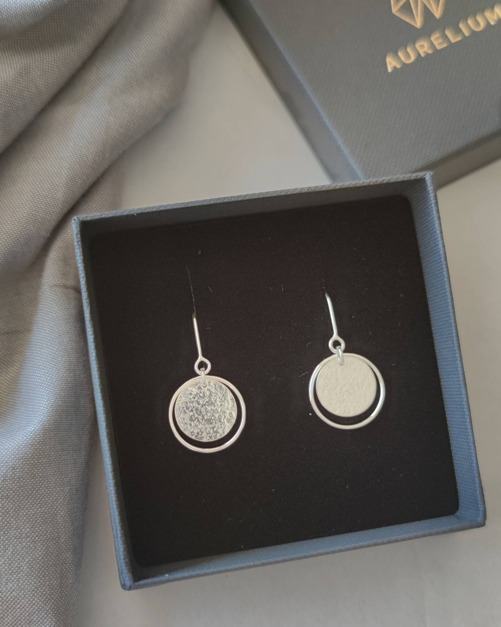 Textured silver circle earrings presented in open black jewellery gift box beside box lid printed with the word "Aurelium" in gold lettering