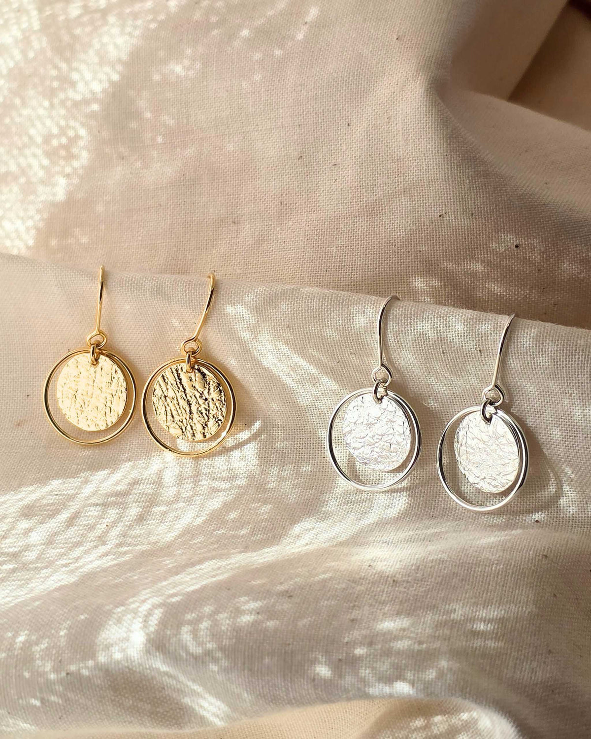 Two pairs of handcrafted gold and silver earrings with textured circles framed within delicate wire loops, hanging from beige fabric