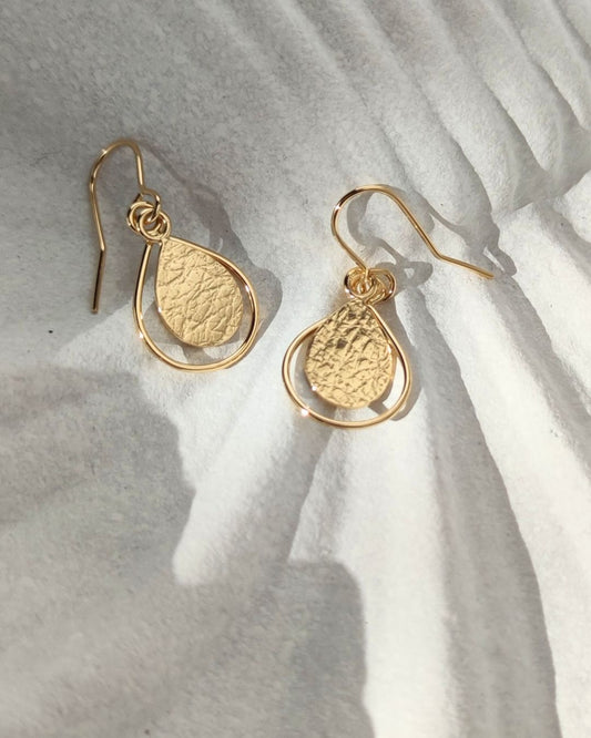 Pair of gold teardrop-shaped earrings with textured surfaces and delicate wire framing, lying on sun-drenched white ceramic shell