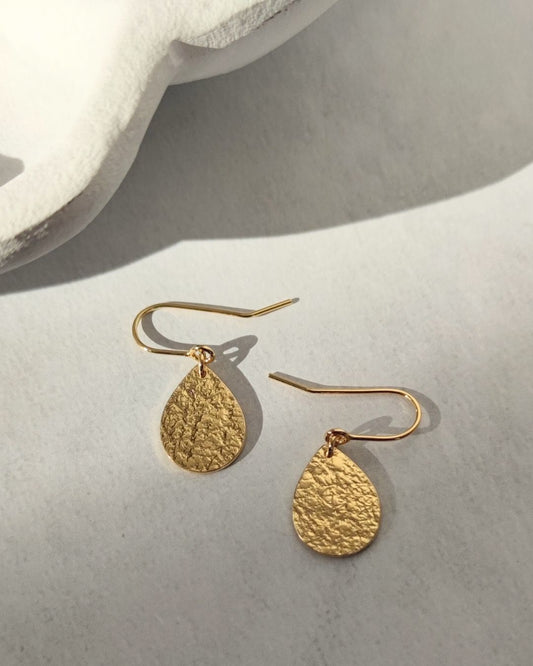 Gold teardrop-shaped textured earrings hanging on handmade gold hooks, lying in sunlight
