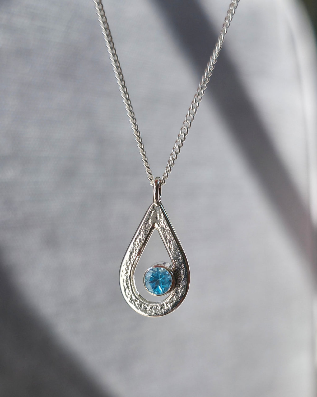 Teardrop-shaped pendant with a blue topaz gemstone on a chain against a grey background