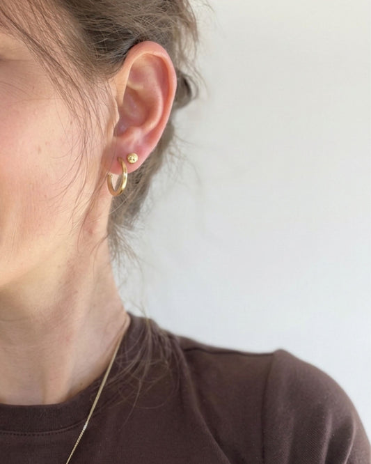 A close-up of a person's ear wearing a small gold hoop earring and a tiny gold stud, against a plain light background