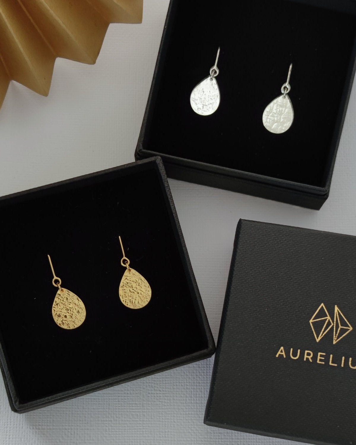 Gold and silver textured teardrop earrings in black gift boxes beside box lid printed with gold logo and "Aurelium" in gold lettering