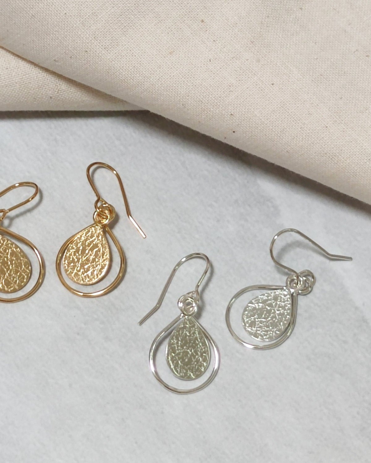 Handmade gold and silver earrings with textured teardrops framed by thin wire teardrops lying beside beige fabric on light coloured surface