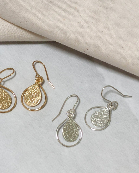 Handmade gold and silver earrings with textured teardrops framed by thin wire teardrops lying beside beige fabric on light coloured surface