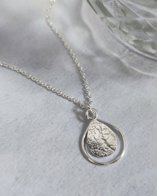 Delicate sterling silver teardrop necklace with textured surface lying on grey marbled surface