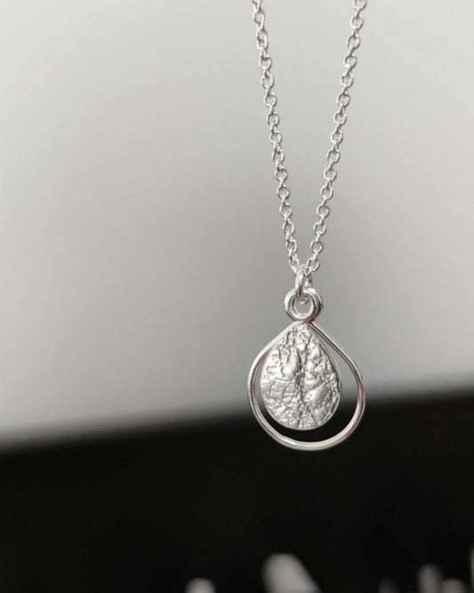 Handmade sterling silver pendant with textured teardrop framed by delicate wire teardrop, hanging from fine silver chain in front of blurred background