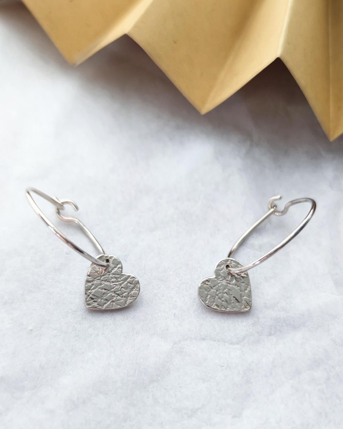 Handmade silver hoop earrings with small textured hearts lying on marbled surface with folded gold paper in background