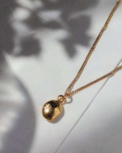 gold molten pebble pendant attached to delicate chain lying in sunshine beside shadow of vase with flowers