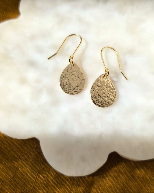 Textured gold teardrop earrings on a white surface lying in diffused sunlight