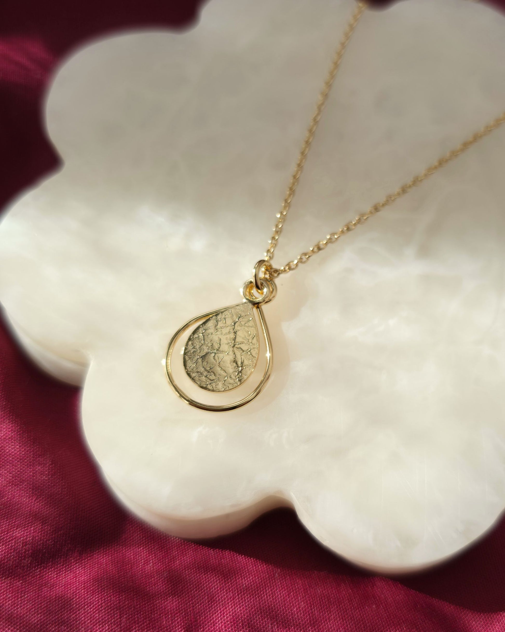 Gold necklace with a teardrop-shaped pendant on a white shell against a red fabric background