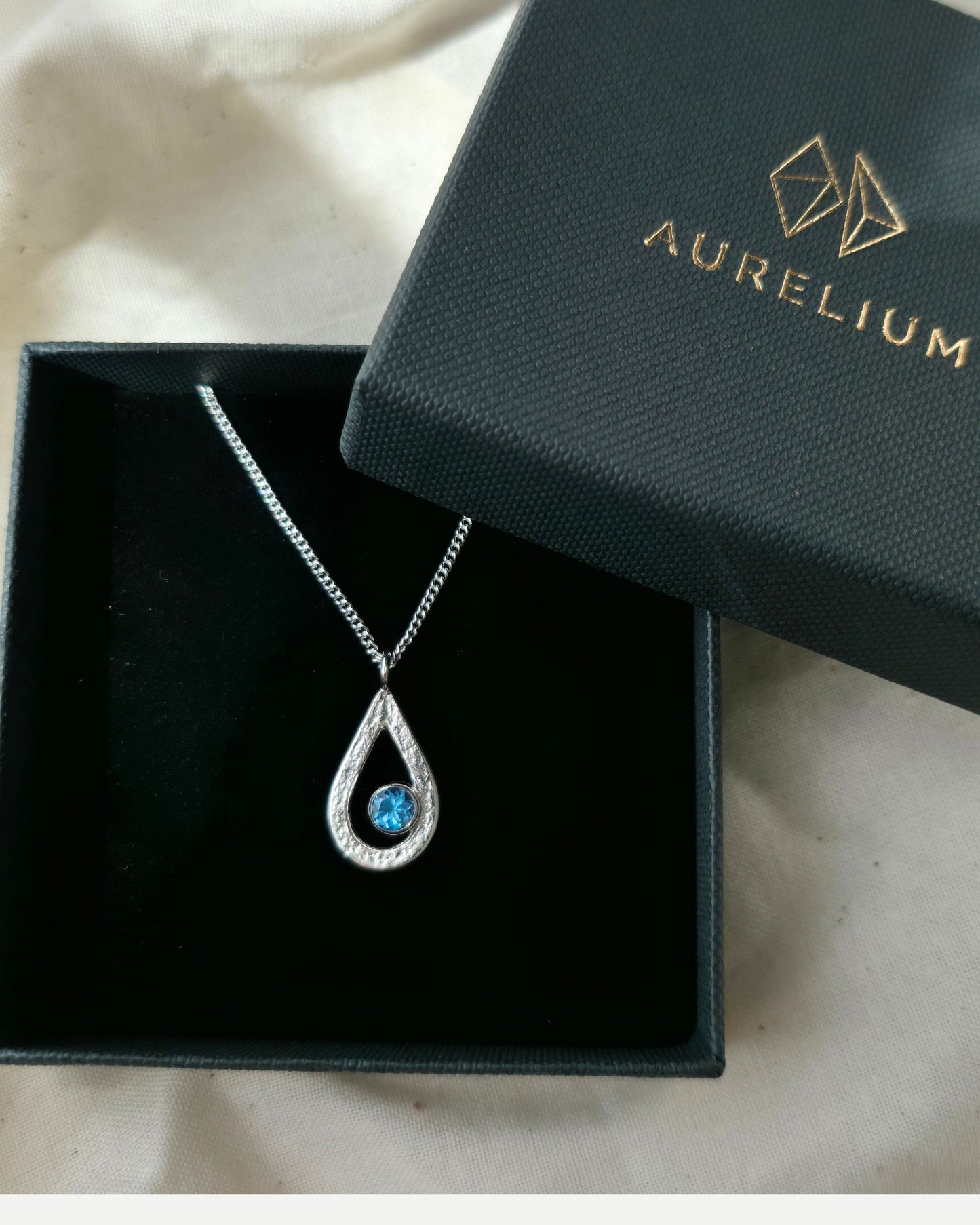 Teardrop-shaped pendant with blue topaz gemstone in a black box with 'Aurelium' branding