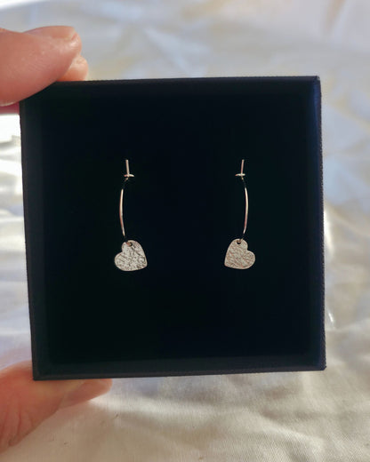 A pair of silver hoop earrings with small textured heart charms, presented in a black gift box held by a hand against a white fabric background