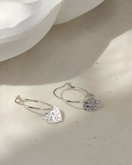 A pair of silver hoop earrings with dangling textured heart charms, displayed on a textured beige fabric surface