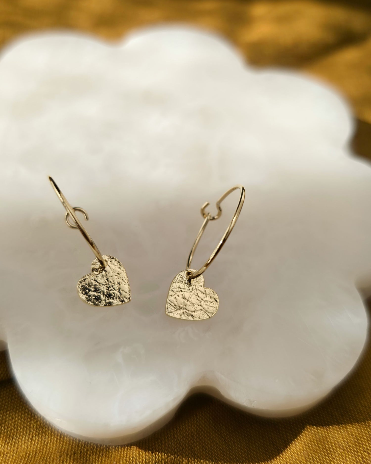 Delicate textured gold heart earrings on cream petal-shaped surface
