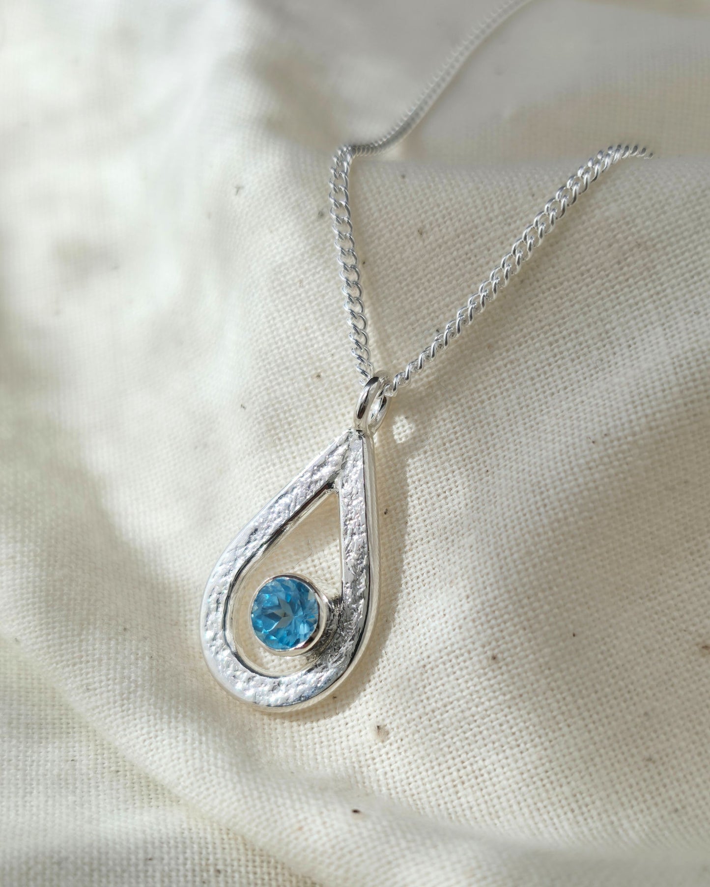 Silver textured teardrop pendant with a blue topaz gemstone on a white fabric background