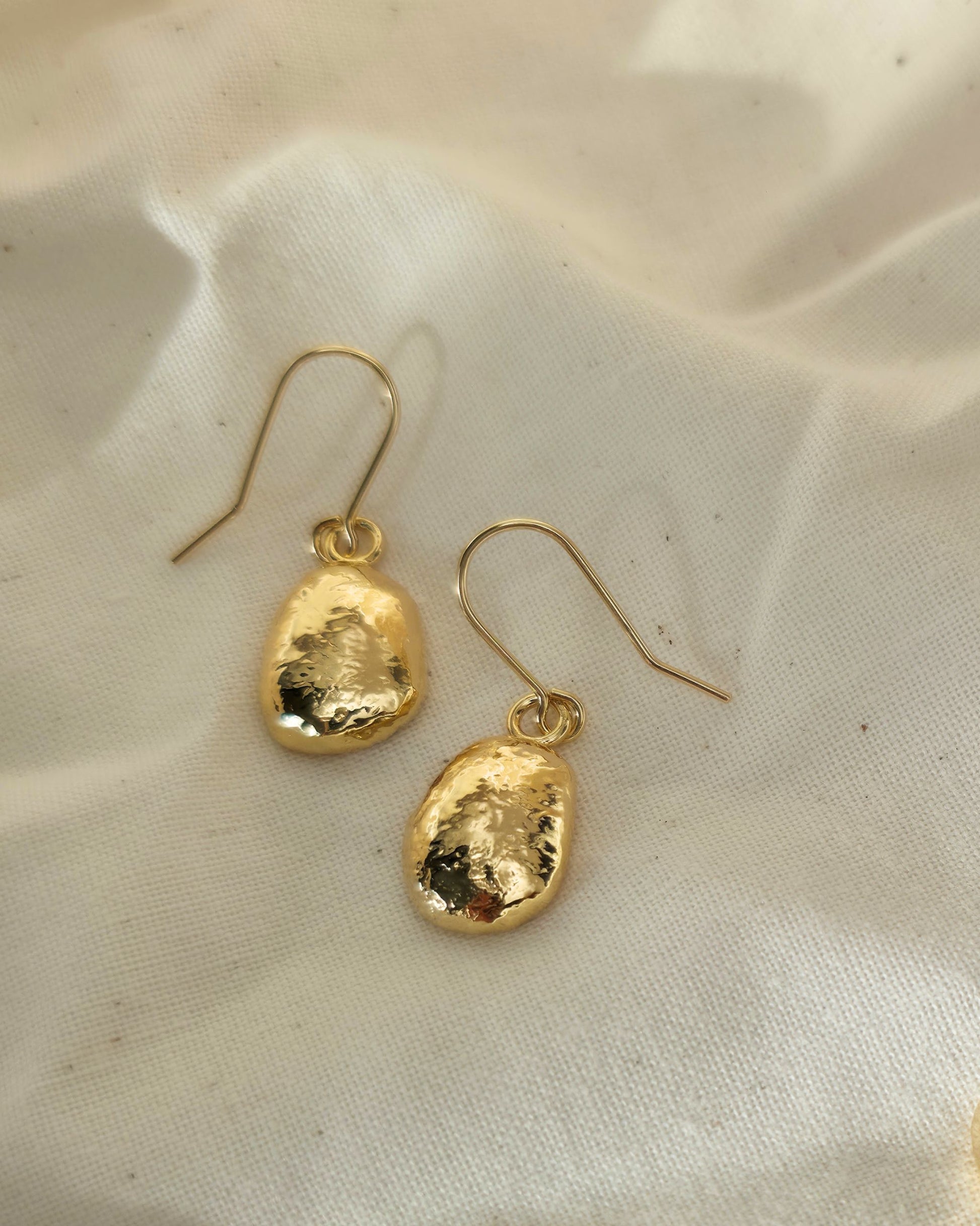 Gold oval shaped earrings with a textured surface lying on beige fabric