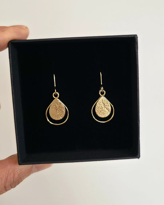 A pair of gold earrings with textured teardrops presented in a black gift box held by a hand