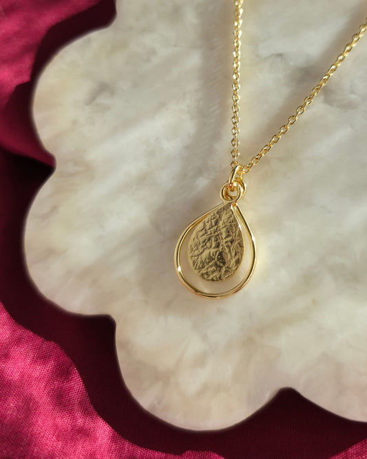 Gold necklace with a textured teardrop-shaped pendant lying in diffused sunlight on a cream surface
