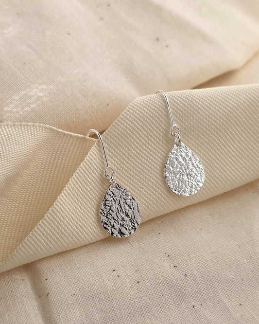 Simple sterling silver teardrop-shaped earrings with a delicate surface texture hanging from thick cream-coloured ribbon
