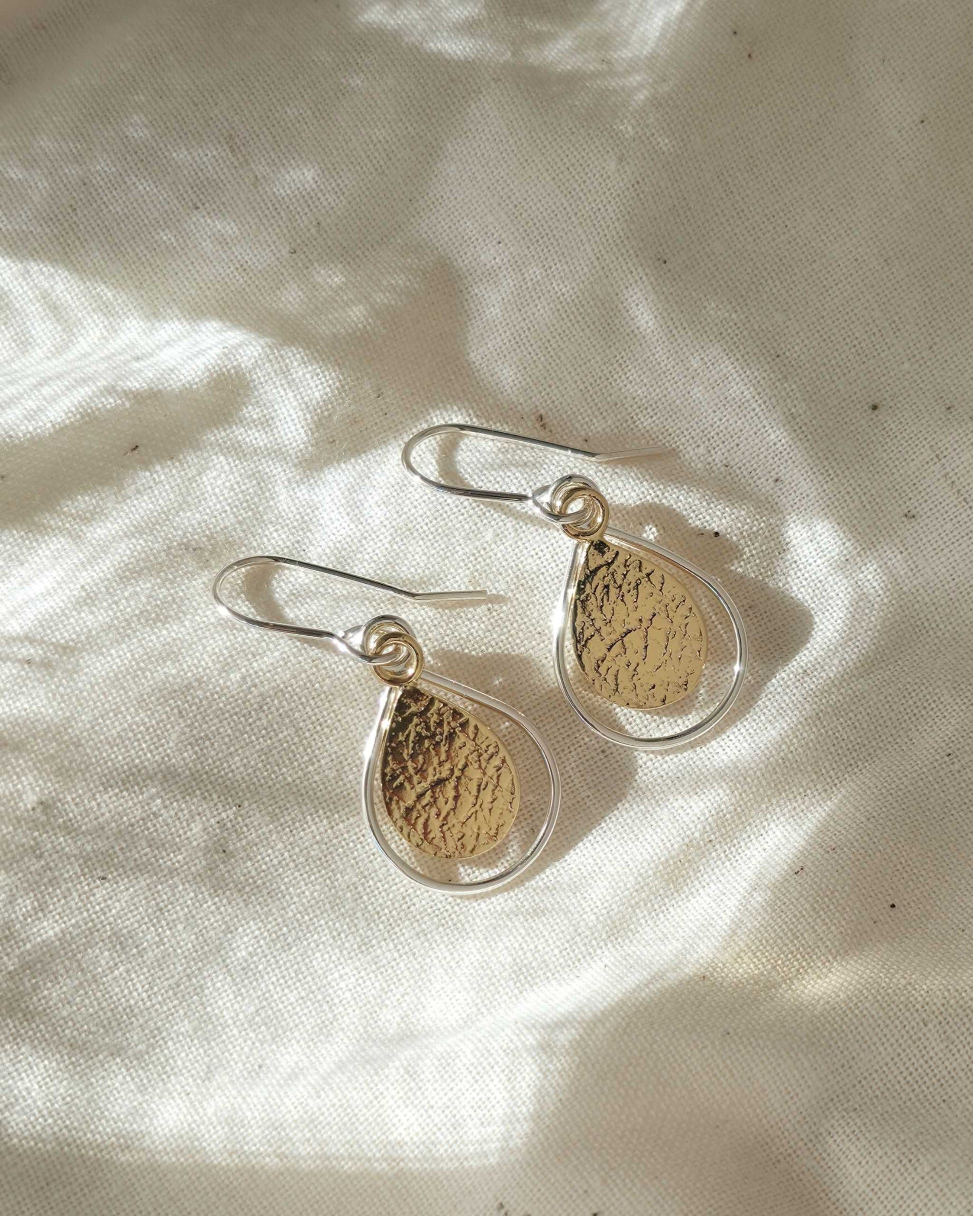 Pair of textured gold teardrop-shaped earrings with silver borders on a light fabric background