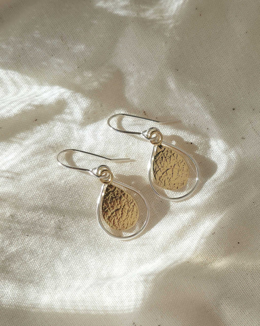 Pair of textured gold teardrop-shaped earrings with silver borders on a light fabric background
