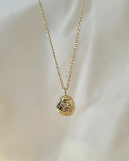 A delicate gold chain necklace with a small, textured gold oval pendant, displayed against a soft, draped white fabric background