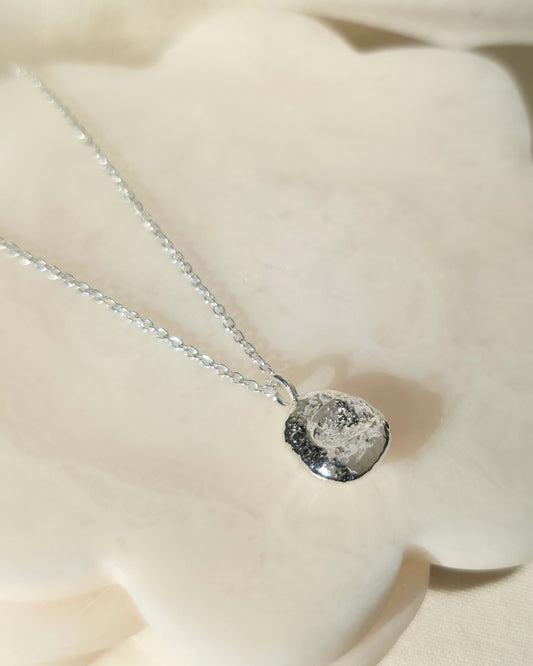 A silver chain necklace with a textured, round silver pendant, displayed on a soft cream-coloured fabric background