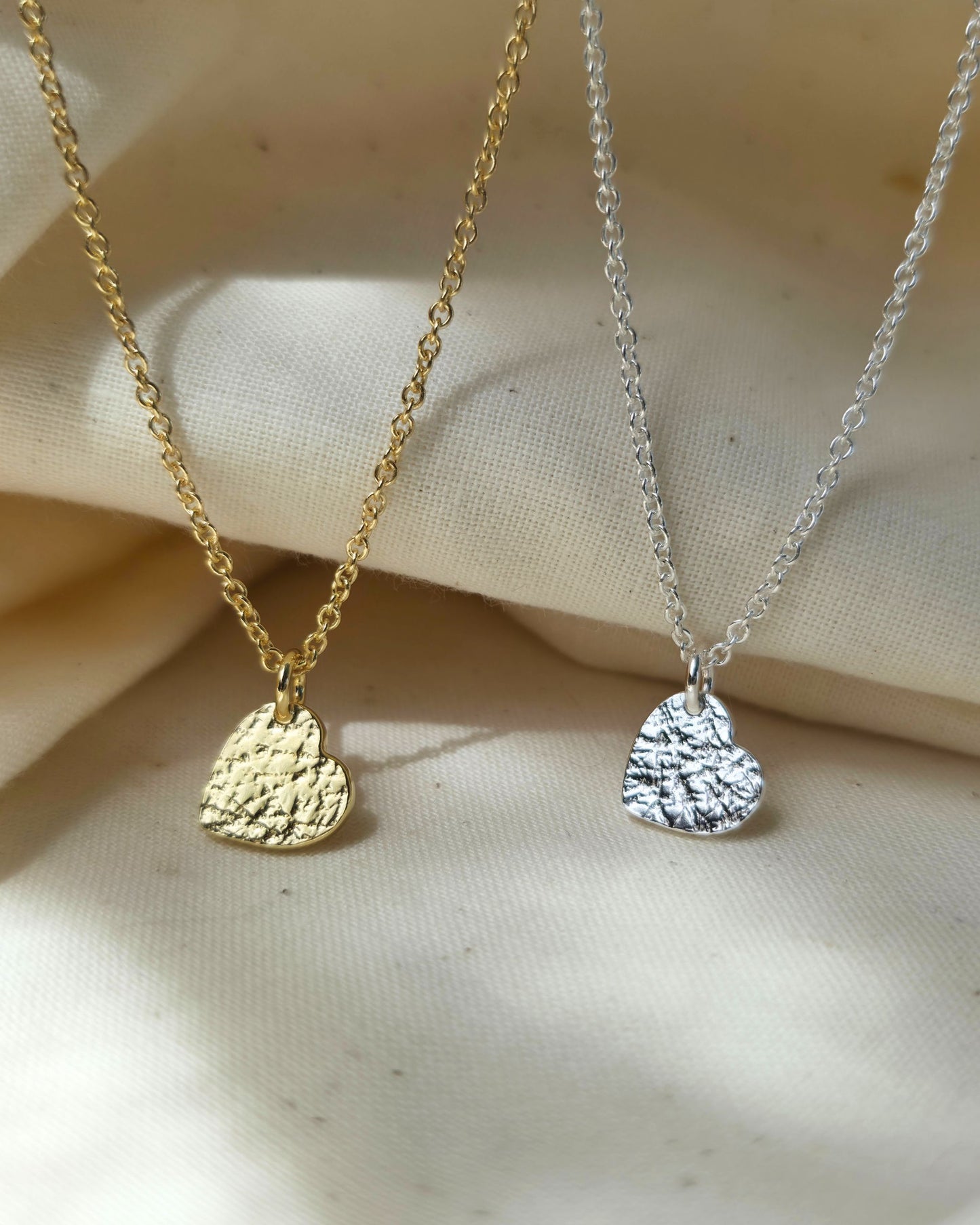 Gold and silver necklaces with textured heart pendants on a beige fabric background
