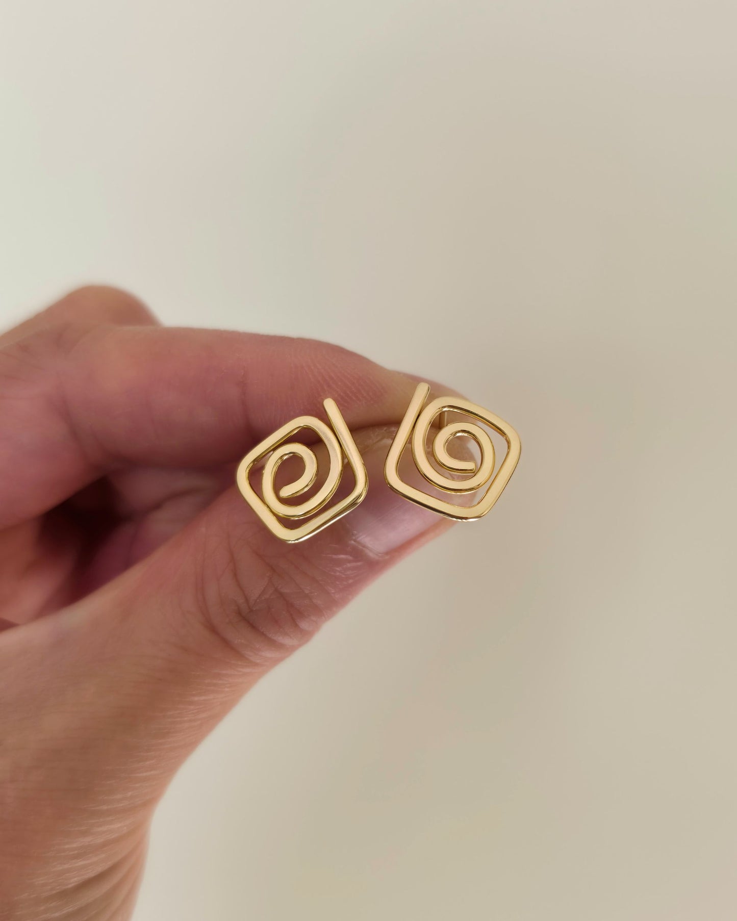 A close-up of a hand holding a pair of gold spiral stud earrings against a plain light beige background.