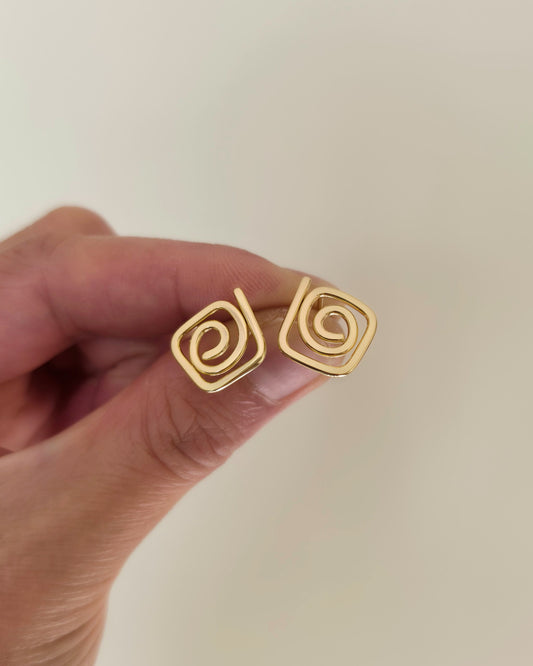 A close-up of a hand holding a pair of gold spiral stud earrings against a plain light beige background.