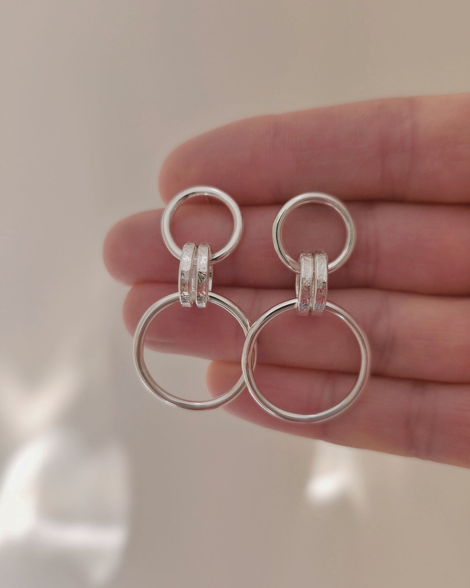A pair of silver loop drop earrings, held in a person's hand against a soft light background