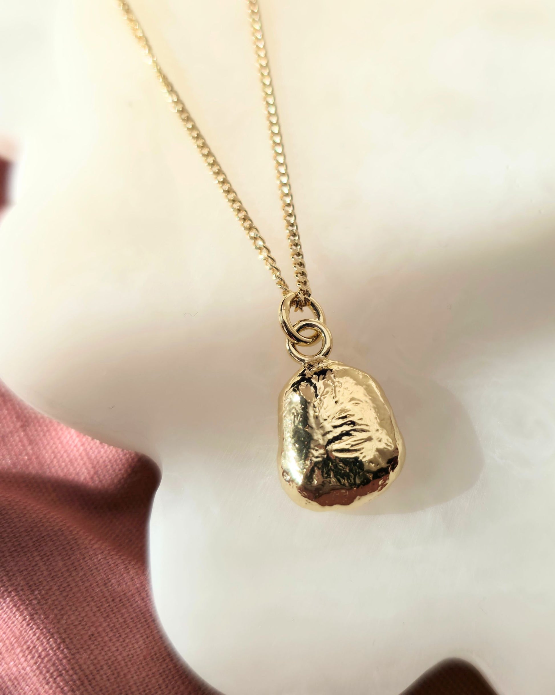 Gold necklace with a textured pendant on a soft pink fabric background