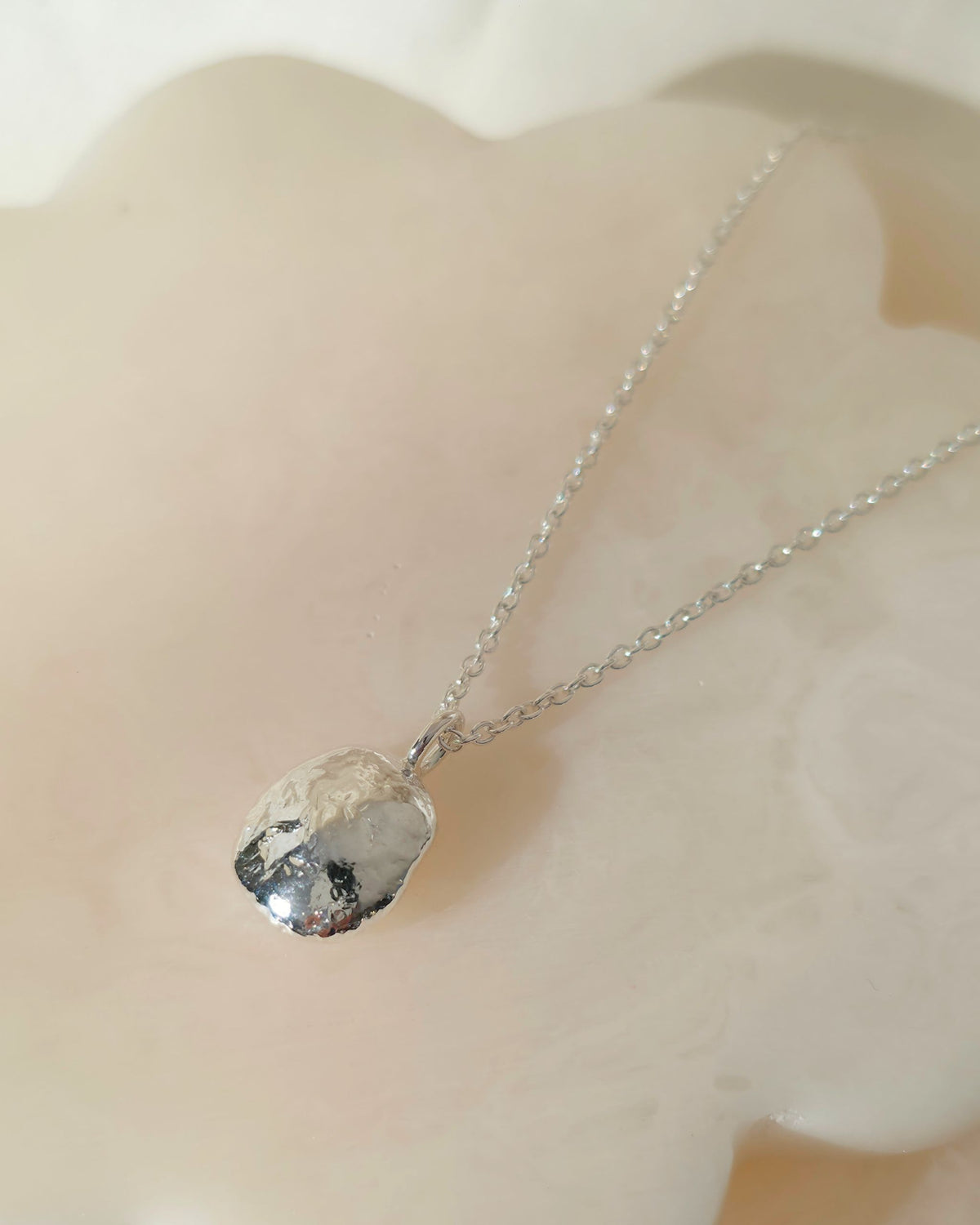 A silver chain necklace with a polished, oval-shaped silver pendant resting on a smooth, pale cream-coloured stone surface