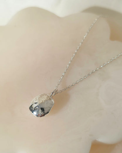 A silver chain necklace with a polished, oval-shaped silver pendant resting on a smooth, pale cream-coloured stone surface