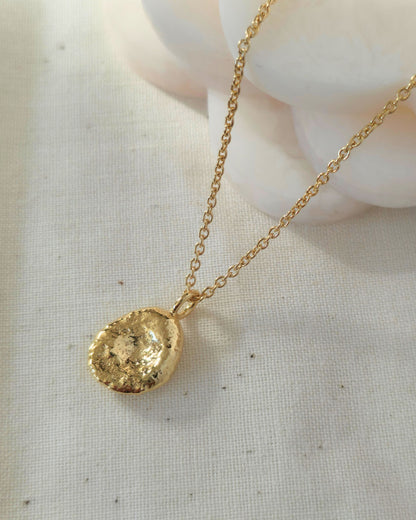 A gold chain necklace with a textured oval-shaped gold pendant, displayed on a light beige linen fabric background